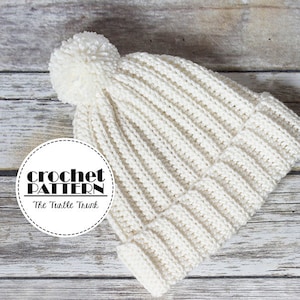 May include: White crocheted beanie with a pom pom on top. The beanie is laying on a wooden surface. The text "crochet pattern" and "The Turtle Trunk" is visible on a white circle.