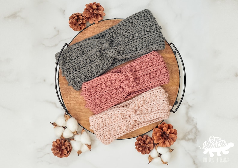 May include: Three crocheted headbands in different colors: gray, pink, and light pink. The headbands are arranged on a wooden tray with a black metal rim. The headbands are all twisted and have a textured pattern.