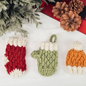 May include: Three crocheted Christmas ornaments: a red and white stocking, a green mitten, and a brown and white hat. The ornaments are on a white surface with pine branches and pine cones in the background.