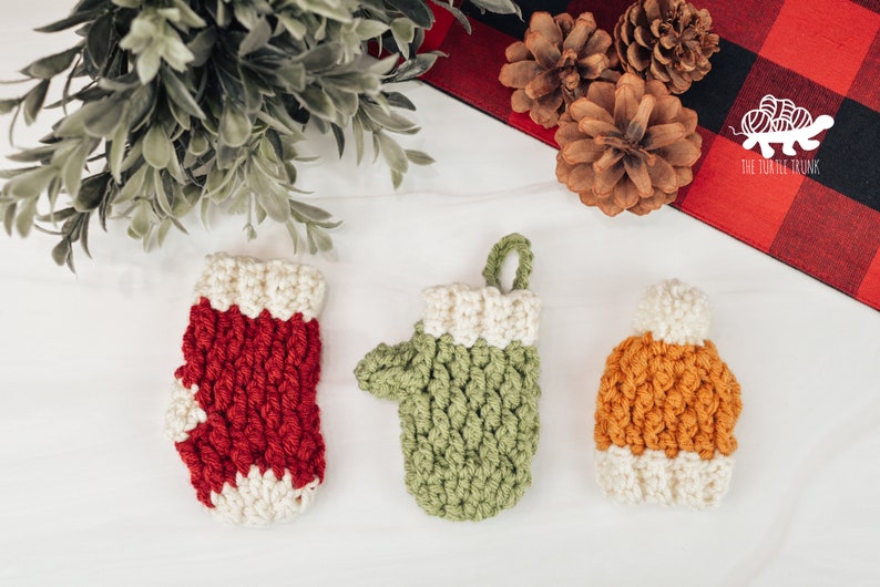 May include: Three crocheted Christmas ornaments: a red and white stocking, a green mitten, and a brown and white hat. The ornaments are on a white surface with pine branches and pine cones in the background.