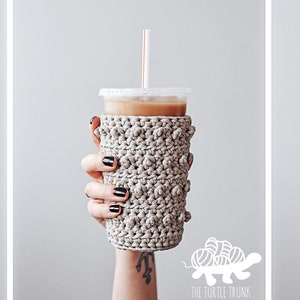May include: A beige crocheted cup cozy with a textured bobble stitch pattern. The cozy is on a clear plastic cup with a straw.
