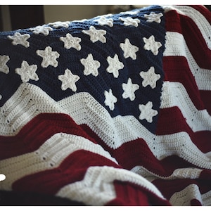 May include: A crocheted American flag blanket in red, white, and blue with white stars on a blue field. The blanket is folded and draped over a chair.