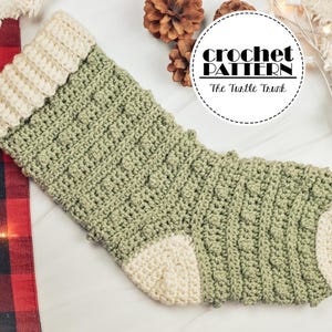May include: A green and white crocheted Christmas stocking with a cable stitch pattern. The stocking is on a white surface with a red and black checked fabric and white fairy lights in the background. The text "crochet pattern" and "The Turtle Trunk" is on a white circle with a black border.
