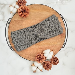 May include: Gray crocheted headband with a twisted knot detail, displayed on a wooden tray with cotton balls and pine cones.