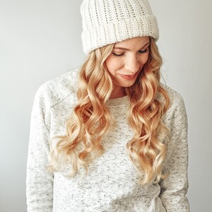 May include: A white crocheted beanie with a fluffy pom pom on top. The beanie has a ribbed texture and is worn by a person with long blonde hair.