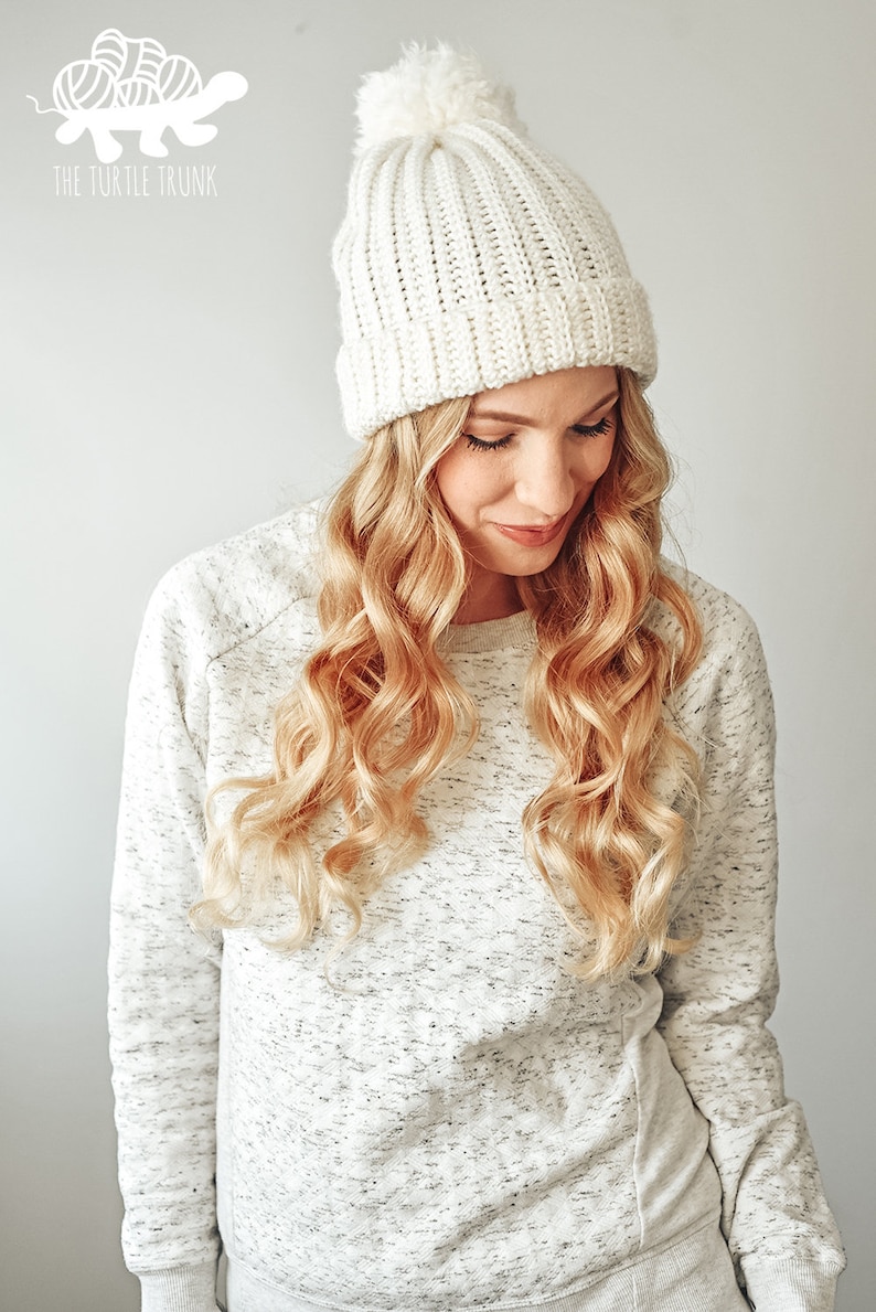 May include: A white crocheted beanie with a fluffy pom pom on top. The beanie has a ribbed texture and is worn by a person with long blonde hair.
