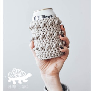 May include: Three crocheted can cozies in a beige color with a textured design. The cozies are on aluminum cans.