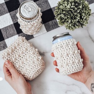 May include: Three crocheted can cozies in a cream color. The cozies are made with a textured stitch and have a small, raised bobble detail. The cozies are shown on a white marble surface. The text "The Turtle Trunk" is visible in the lower left corner of the image.
