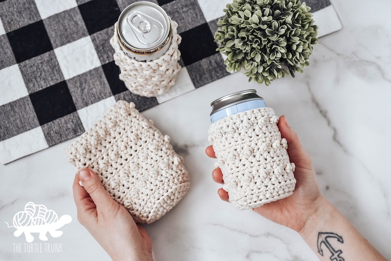 May include: Three crocheted can cozies in a cream color. The cozies are made with a textured stitch and have a small, raised bobble detail. The cozies are shown on a white marble surface. The text "The Turtle Trunk" is visible in the lower left corner of the image.