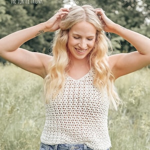 May include: A woman wearing a white crochet tank top with a v-neck and a pair of light blue denim shorts. She is standing in a field of tall grass.