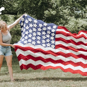 May include: A woman holds up a large crocheted American flag blanket. The blanket is red, white, and blue with a wavy design. The stars are white and the stripes are red and white.