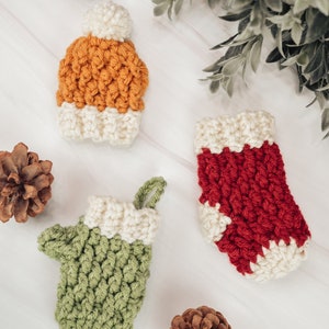 May include: Three crocheted ornaments in the shape of a stocking, a mitten, and a hat. The stocking is red with white trim, the mitten is green with white trim, and the hat is orange with white trim. The ornaments are on a white surface with pine cones.