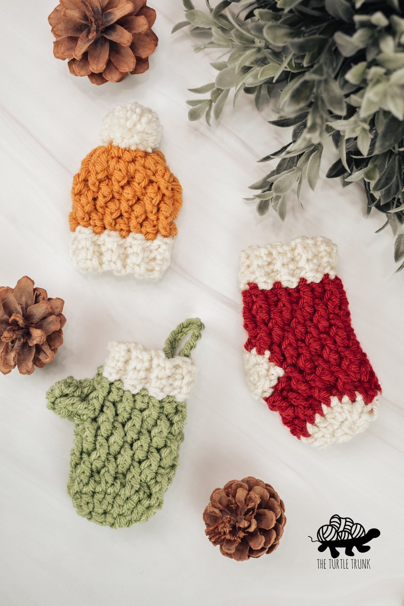 May include: Three crocheted ornaments in the shape of a stocking, a mitten, and a hat. The stocking is red with white trim, the mitten is green with white trim, and the hat is orange with white trim. The ornaments are on a white surface with pine cones.