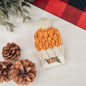 May include: A crocheted orange and white beanie with a white pom pom on top. The beanie is sitting on a white surface with three pine cones.