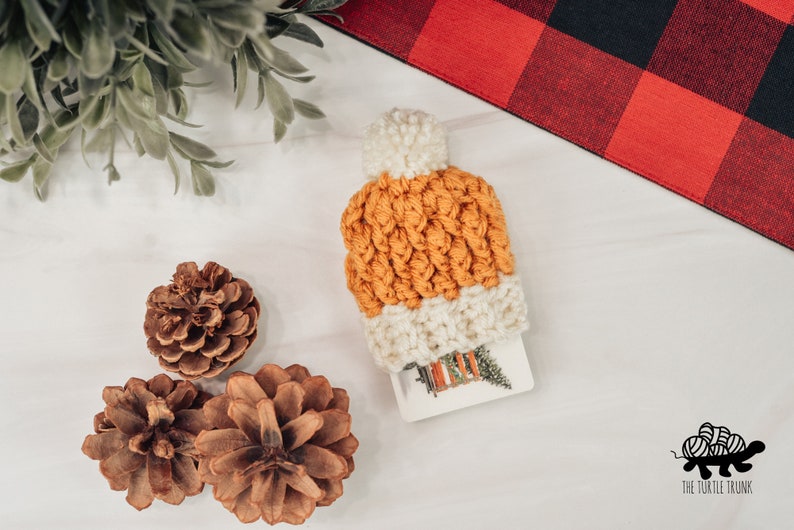 May include: A crocheted orange and white beanie with a white pom pom on top. The beanie is sitting on a white surface with three pine cones.