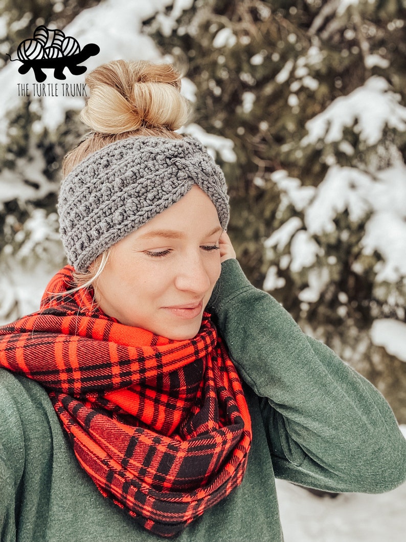 May include: A woman wearing a gray crocheted headband and a red and black plaid scarf. The background is a snowy forest.