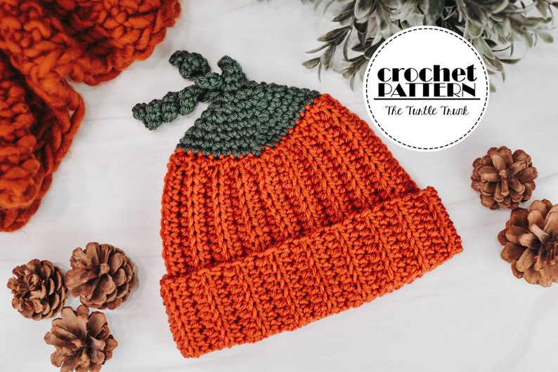 A ribbed, crochet Pumpkin Beanie laying on a white surface with a scarf, pine cones, and plant around it.