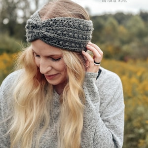 May include: A gray crocheted headband with a twisted knot detail. The headband is worn by a person with long blonde hair. The background is a blurred image of a field of yellow flowers.