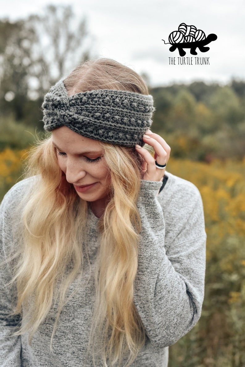 May include: A gray crocheted headband with a twisted knot detail. The headband is worn by a person with long blonde hair. The background is a blurred image of a field of yellow flowers.