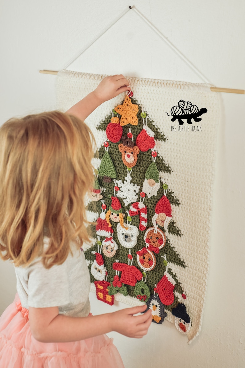 Puede incluir: Un ni&ntilde;o cuelga un adorno de ping&uuml;ino de crochet en un calendario de adviento de &aacute;rbol de Navidad de crochet blanco y verde. El calendario est&aacute; decorado con varios adornos de crochet, incluyendo una estrella, un reno, un mu&ntilde;eco de nieve y un hombre de jengibre. El texto "THE TURTLE TRUNK" es visible en la esquina inferior derecha.