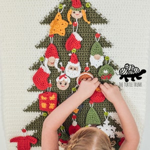 Puede incluir: Un ni&ntilde;o decora un &aacute;rbol de Navidad de crochet con varios adornos, incluyendo un gnomo, una estrella, una media, un regalo y un Pap&aacute; Noel. El &aacute;rbol es verde y blanco, y los adornos son rojos, verdes y blancos. El texto "THE TURTLE TRUNK" es visible en la esquina inferior derecha de la imagen.