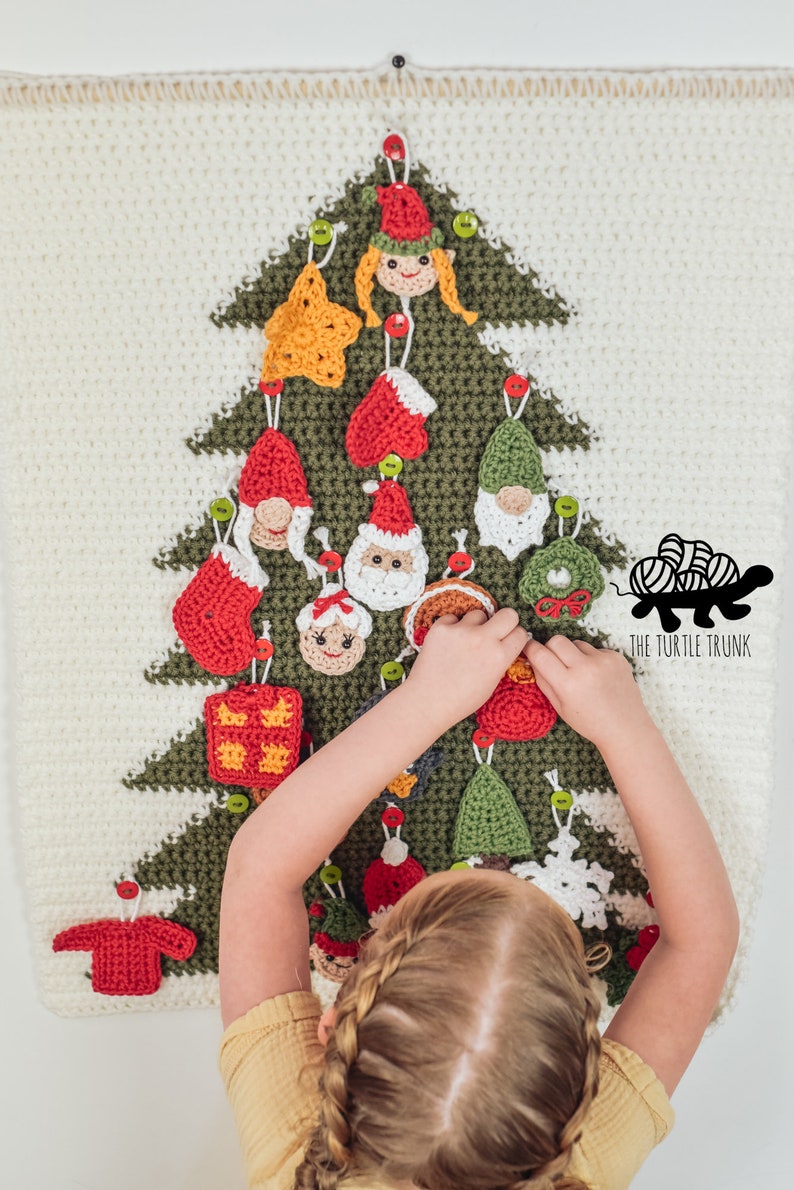 Puede incluir: Un ni&ntilde;o decora un &aacute;rbol de Navidad de crochet con varios adornos, incluyendo un gnomo, una estrella, una media, un regalo y un Pap&aacute; Noel. El &aacute;rbol es verde y blanco, y los adornos son rojos, verdes y blancos. El texto "THE TURTLE TRUNK" es visible en la esquina inferior derecha de la imagen.