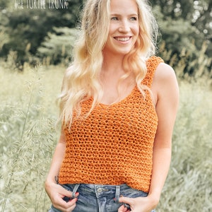 May include: A woman wearing a crocheted orange tank top with a v-neck and a pair of blue denim shorts. The top is a loose fit and has a textured pattern. The woman is standing in a field of tall grass.