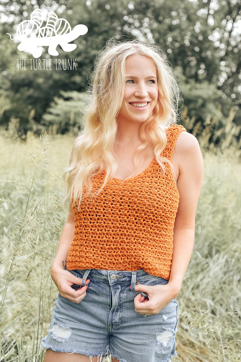 May include: A woman wearing a crocheted orange tank top with a v-neck and a pair of blue denim shorts. The top is a loose fit and has a textured pattern. The woman is standing in a field of tall grass.