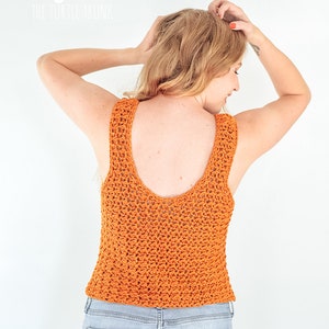 May include: A woman wearing a burnt orange crochet tank top with a deep V-neck back. The tank top is made with a simple stitch pattern and has a relaxed fit. The woman is wearing light blue jeans.