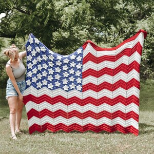 May include: A woman holds up a large crocheted American flag blanket with red, white, and blue stripes and stars. The blanket is made with a chevron pattern. The woman is standing in a grassy field.
