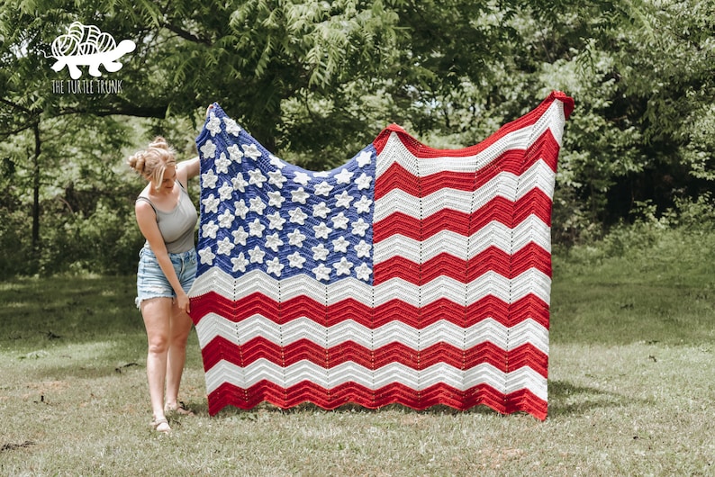 May include: A woman holds up a large crocheted American flag blanket with red, white, and blue stripes and stars. The blanket is made with a chevron pattern. The woman is standing in a grassy field.