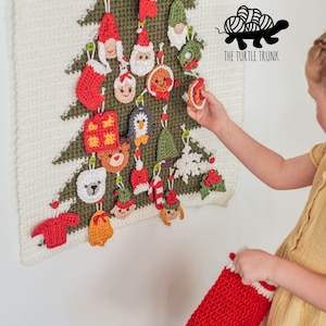 Puede incluir: Un calendario de adviento de Navidad tejido a crochet en forma de &aacute;rbol con una variedad de adornos coloridos, que incluyen un ping&uuml;ino, un mu&ntilde;eco de nieve, un hombre de jengibre y una media. Un ni&ntilde;o sostiene una media roja tejida a crochet.