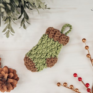May include: A small green and brown crocheted stocking with a loop for hanging. The stocking is on a white surface with pine cones and a sprig of berries.