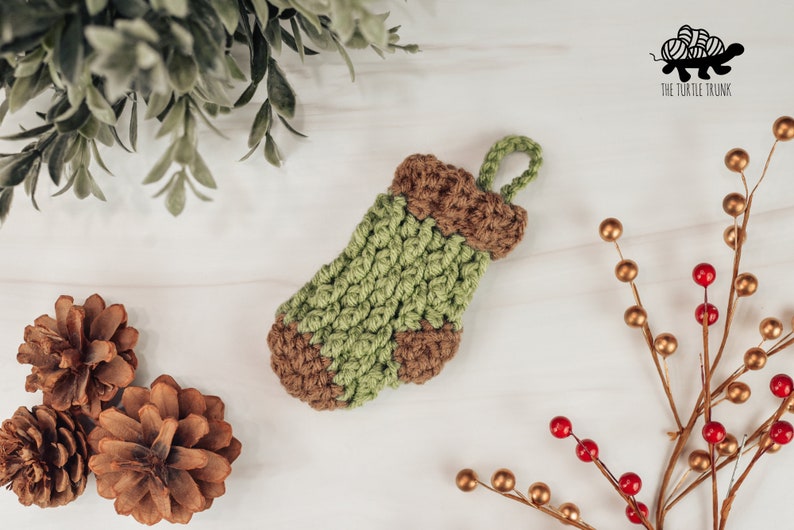 May include: A small green and brown crocheted stocking with a loop for hanging. The stocking is on a white surface with pine cones and a sprig of berries.