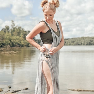 May include: A woman wearing a long, grey crochet dress with a high slit on the side. The dress is made with a simple stitch pattern and has a relaxed fit. The woman is standing on a beach with a body of water in the background.