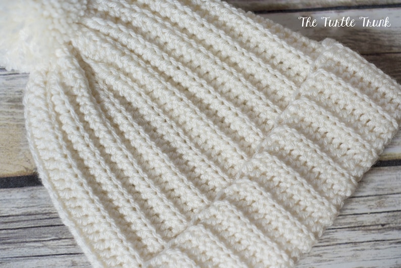 May include: A white crocheted beanie with a fluffy pom pom on top. The beanie has a ribbed texture and is made with a soft yarn.