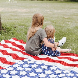 May include: Two children sit on a red, white, and blue crocheted blanket with a chevron pattern. The blanket has a blue section with white stars. The text "THE TURTLE TRUNK" is visible in the upper left corner of the image.