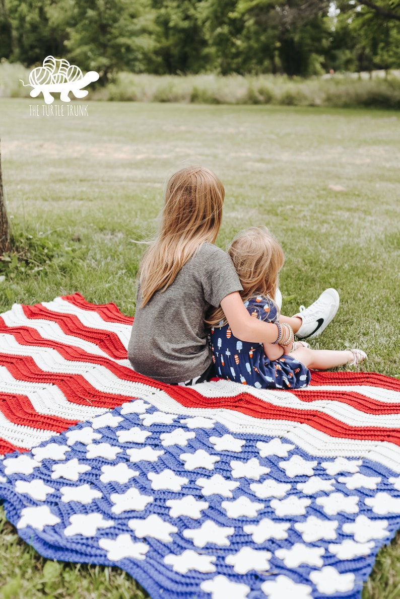 May include: Two children sit on a red, white, and blue crocheted blanket with a chevron pattern. The blanket has a blue section with white stars. The text "THE TURTLE TRUNK" is visible in the upper left corner of the image.