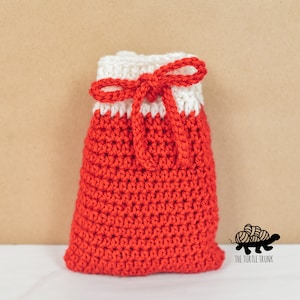 Puede incluir: Una bolsa de cord&oacute;n de crochet roja y blanca con un lazo blanco. La bolsa est&aacute; sobre una superficie blanca con un fondo marr&oacute;n. El texto "The Turtle Trunk" es visible en la esquina inferior derecha de la imagen.