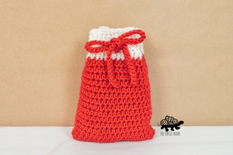 Puede incluir: Una bolsa de cord&oacute;n de crochet roja y blanca con un lazo blanco. La bolsa est&aacute; sobre una superficie blanca con un fondo marr&oacute;n. El texto "The Turtle Trunk" es visible en la esquina inferior derecha de la imagen.