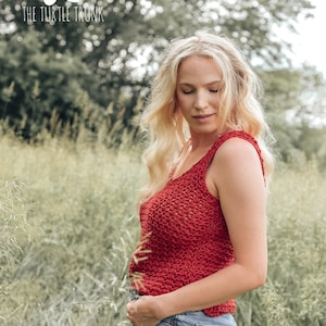 May include: A woman wearing a red crochet tank top and denim shorts stands in a field of tall grass. The tank top has a loose, open weave. The woman is looking down at her hands.