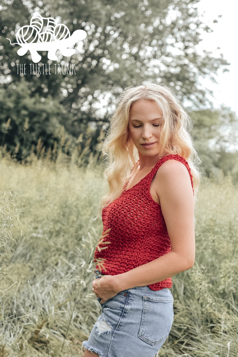 May include: A woman wearing a red crochet tank top and denim shorts stands in a field of tall grass. The tank top has a loose, open weave. The woman is looking down at her hands.