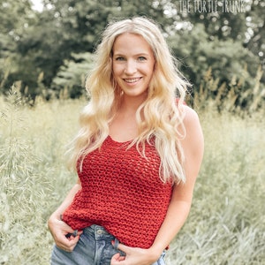 May include: A woman wearing a red crochet tank top with a crocheted stitch pattern. She is standing in a field of tall grass.