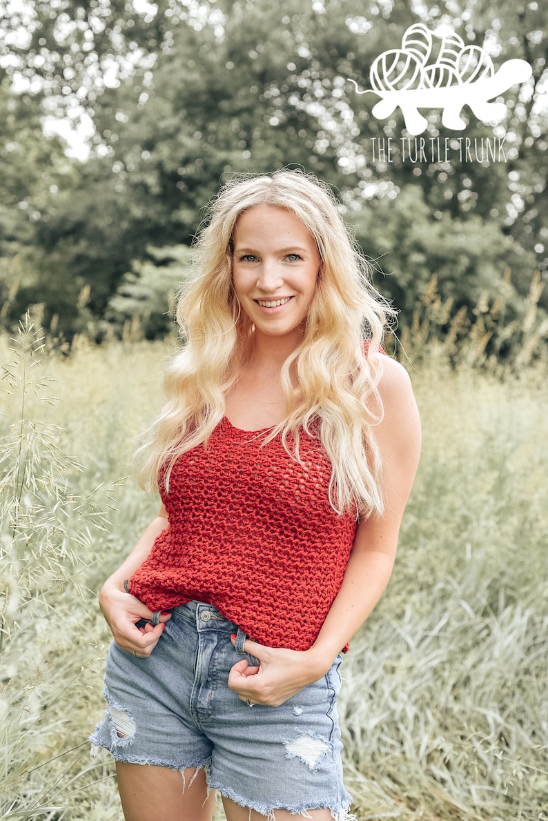 May include: A woman wearing a red crochet tank top with a crocheted stitch pattern. She is standing in a field of tall grass.