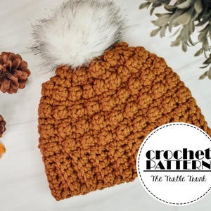 May include: A handmade, textured, crochet hat in a warm, golden-brown colour, topped with a fluffy white pom-pom. The hat is surrounded by pinecones and autumn leaves. The image includes a circular logo that says "crochet PATTERN The Turtle Trunk."