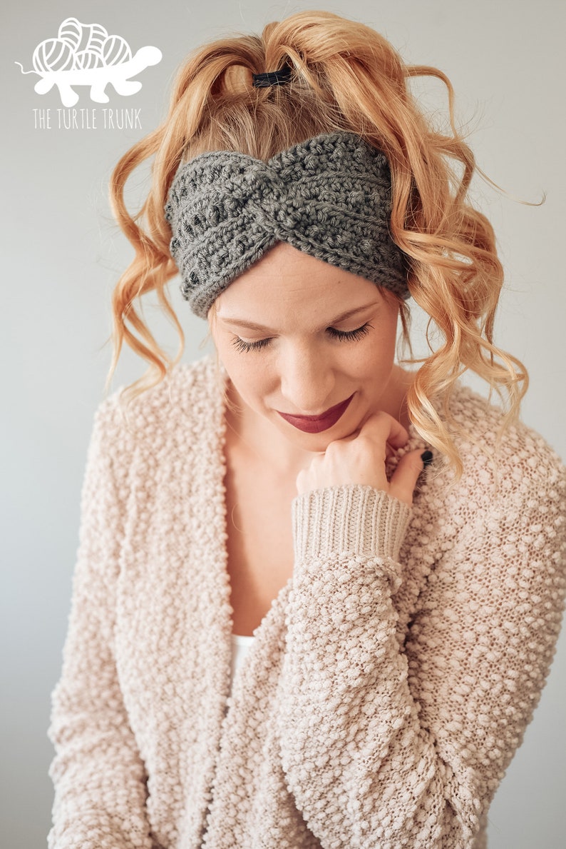 May include: A woman with long, curly blonde hair is wearing a gray crocheted headband. The headband is twisted and has a textured design. The woman is wearing a light beige cardigan with a textured pattern.