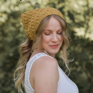 Puede incluir: Una mujer lleva una boina de crochet de color mostaza con un nudo. La boina tiene un patr&oacute;n de punto texturizado. La mujer lleva un top blanco sin mangas con un ribete de encaje.