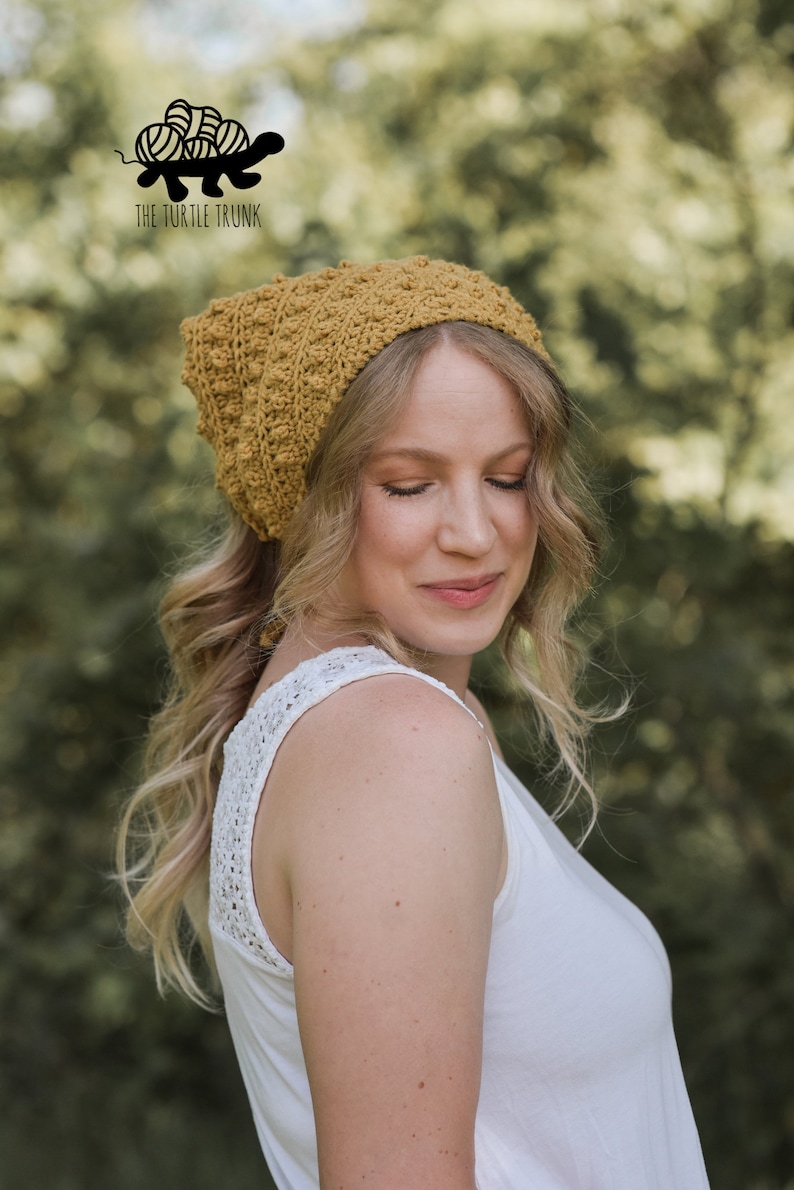Puede incluir: Una mujer lleva una boina de crochet de color mostaza con un nudo. La boina tiene un patr&oacute;n de punto texturizado. La mujer lleva un top blanco sin mangas con un ribete de encaje.