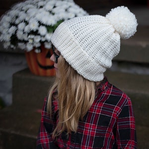 May include: A white crocheted beanie with a white pom pom on top. The beanie is worn by a person with long blonde hair and a red and black plaid shirt.