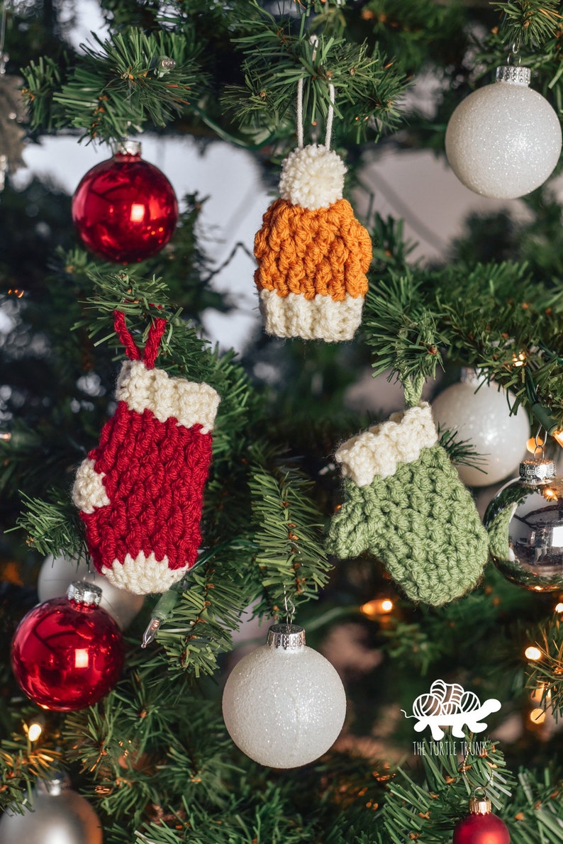 May include: A Christmas tree decorated with crocheted ornaments, including a red mitten, a green mitten, and an orange and white hat. The tree is also decorated with red and white ornaments.